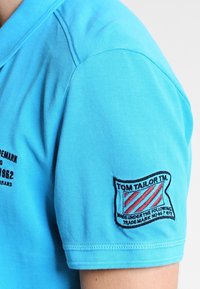 Polo léger bleu en coton texturé à manches courtes, avec un patch logo et du texte sur la manche gauche.