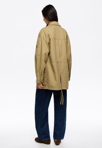 Chaqueta de grandes dimensiones en color caqui con una abertura en la parte trasera, dos bolsillos laterales y mangas largas. Combinada con pantalones vaqueros de pierna ancha en azul y calzado marrón.