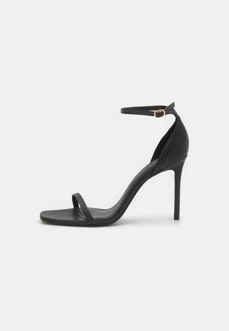 Sandale noire à talon haut avec fine bride sur les orteils et bride à la cheville avec petite boucle dorée sur fond blanc.