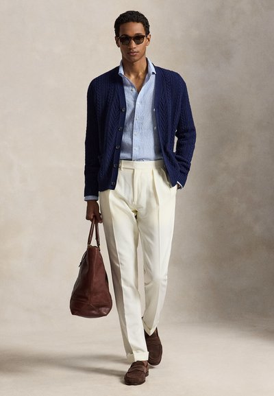 Homme portant un cardigan bleu marine, une chemise bleu clair, un pantalon crème, des mocassins marron, des lunettes de soleil, tenant un sac en cuir marron, debout devant un fond neutre.