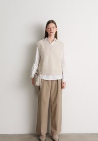 Suéter sem mangas tricotado em bege sobre uma camisa branca, combinado com calças largas em bege. Apresenta um decote em V e um padrão texturizado.