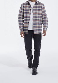 Man loopt in een blauw, oranje en witte geruite shirt over een wit t-shirt, zwarte broek en zwarte schoenen, tegen een witte achtergrond.