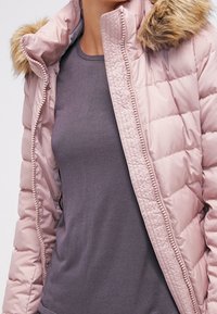 Ljusrosa pufferjacka med huva i faux päls, dragkedjestängning och kviltad textur. Bärs över en grå figurnära topp.