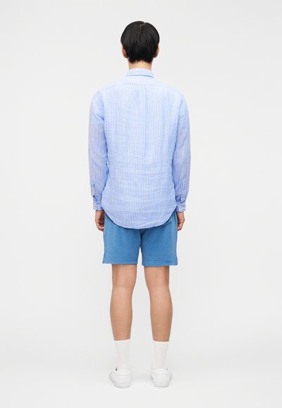 Άνδρας που φοράει μπλε και λευκό ριγέ μακρυμάνικο πουκάμισο, μπλε σορτς, λευκές κάλτσες και λευκά αθλητικά παπούτσια, στέκεται γυρισμένος με την πλάτη σε λευκό φόντο.