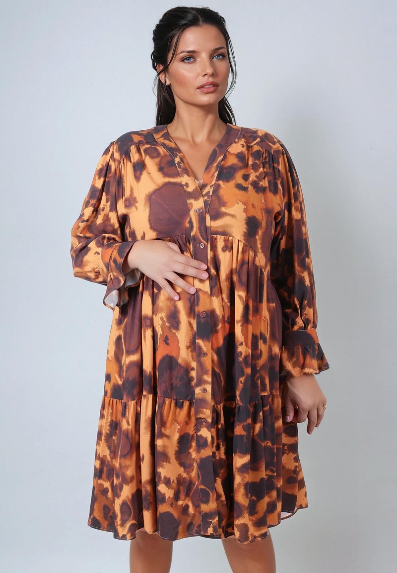 Femme portant une robe longue à manches longues, boutonnée et ample, avec des motifs tie-dye orange et brun foncé, posant devant un fond gris clair uni.