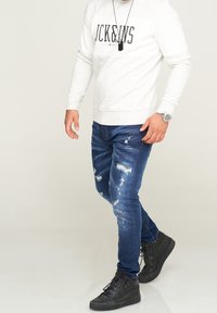 Hombre vestido con sudadera blanca, jeans azules desgastados con manchas de pintura, zapatillas altas negras, reloj plateado y collar con colgante negro.