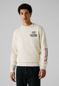 Crème sweatshirt met ronde hals, met het "RED BULL RACING" logo op de borst en "Authentic Race Gear" op de mouw. De stof is glad.