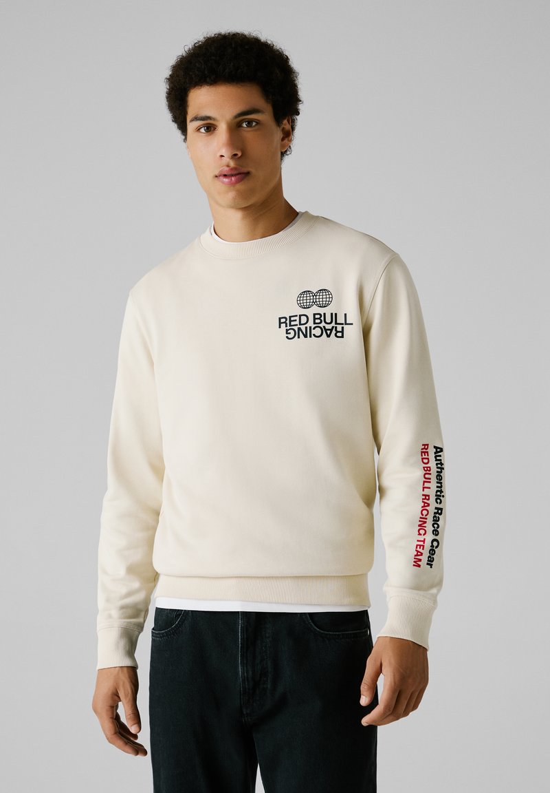 Crème sweatshirt met ronde hals, met het "RED BULL RACING" logo op de borst en "Authentic Race Gear" op de mouw. De stof is glad.