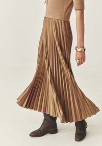 Falda midi con pliegues en suave tela marrón, que presenta una cintura alta y un diseño fluido. Combinada con botas marrones hasta la rodilla y accesorios dorados.