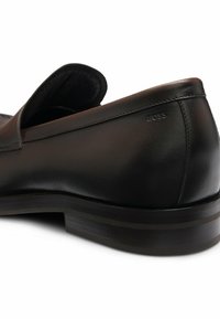 Scarpa nera in pelle slip-on con finitura liscia, punta arrotondata e un discreto logo sul tacco. Dotata di una suola bassa e testurizzata.