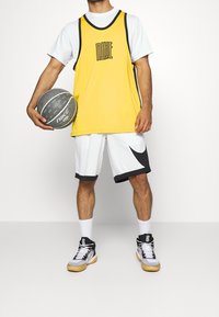 Maillot de basket-ball jaune avec des accents noirs, associé à un short blanc arborant un logo Nike noir. Le modèle tient un ballon de basket gris, portant des baskets noires.