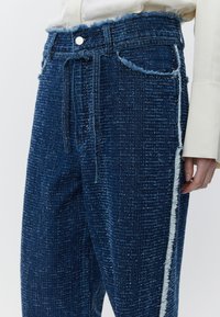Niebieskie spodnie dżinsowe z fakturowanym wykończeniem, wyposażone w postrzępiony pas i boczny szew. Detale obejmują zapięcie na guziki i sznurek do ściągania.