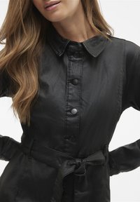 Czarny płaszcz z paskiem, wykonany z gładkiego, błyszczącego materiału. Posiada spiczasty kołnierz, zapięcie na guziki oraz długie rękawy. Teksturowana tkanina.