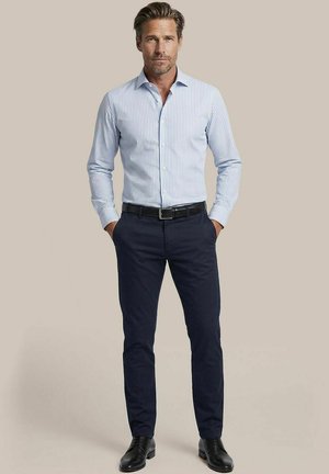 Man draagt een lichtblauw gestreept overhemd, donkerblauwe broek, zwarte riem en zwarte nette schoenen, staat met handen in de zakken.