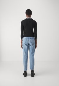 Haut noir à manches longues avec col blanc et fermeture éclair au dos, associé à un jean droit bleu clair. Des bottes noires à semelle épaisse complètent le look.