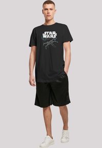 Svart bomull T-skjorte med hvit "STAR WARS" tekst og et grafisk design av en romfighter. Modell iført svarte shorts og hvite joggesko.