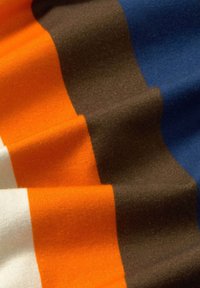 Stribet stof med orange, hvid, brun og blå sektioner. Glat tekstur med ensartet strikkemønster og synlige syninger.