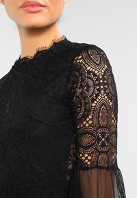 Robe en dentelle noire ornée de motifs floraux complexes, avec des manches en tulle transparent, un col haut et de délicates bordures texturées.