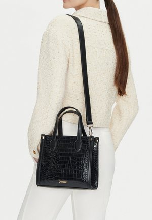 Mujer con cabello largo y castaño, que lleva una chaqueta color crema y pantalones blancos, y lleva un bolso negro con patrón de cocodrilo y correa para el hombro.