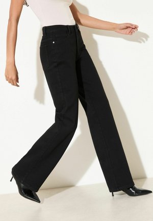 Osoba nosząca czarne dżinsy z wysokim stanem o szerokich nogawkach oraz czarne buty na obcasie z zakończeniem w szpic, idąca na białym tle.