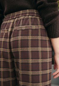 Pantaloni a quadri marroni con tessuto morbido, vita elasticizzata e due tasche posteriori. Il motivo presenta sottili linee gialle lungo tutto il tessuto.