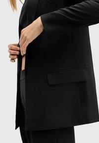 Blazer nero su misura con rever a pistagna, taschino sul petto e due tasche laterali. Tessuto morbido con una texture liscia, design leggermente sagomato.