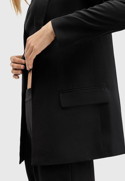 Blazer noir ajusté avec une texture lisse et une poche latérale unique. Le design présente une silhouette cintrée et un revers cranté.