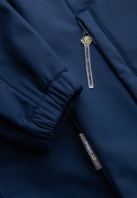 Gros plan du tissu d'une veste bleu marine montrant un poignet élastique et deux curseurs de fermeture éclair étiquetés "ombro performance."