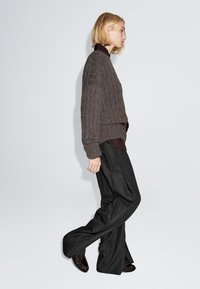 Brun ribbet strikket sweater med sænkede skuldre, lagd oven på en mørk skjorte, parret med brede sorte ternede bukser og mørke sko.