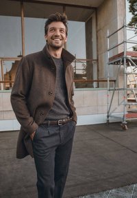 Brun ullkappa med hög krage, grå stickad tröja under, mörka skräddarsydda byxor och ett flätat bälte. Ler i en stadsmiljö.