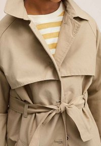 Beżowy płaszcz trench z szerokim kołnierzem i paskiem do wiązania, z podwójnym zapięciem i teksturowanym materiałem. Pod spodem biało-żółty sweter w paski.