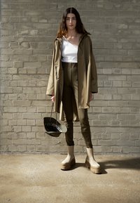 Cămașă impermeabilă bej peste un top alb, pantaloni ajustați verde măsliniu și cizme groase bej. Geantă neagră cu interior verde. Fundal cu textură de cărămidă.