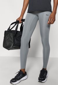 Άτομο που φοράει γκρι κολάν Under Armour και μαύρα αθλητικά παπούτσια, κρατώντας μια μαύρη σακούλα τύπου duffel στο πλάι του, μπροστά σε απλό φόντο.