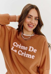 Hnědá mikina, oversized střih, na přední straně s růžovým texturovaným nápisem "Crème De La Crème". Viditelný náhrdelník z vícebarevných korálků.