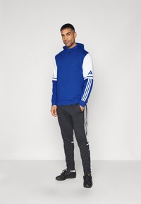 Blauer Hoodie mit weißen Ärmeln, der drei Streifen aufweist, kombiniert mit schwarzen Jogginghosen mit weißen Seitenstreifen und schwarzen Sportschuhen.