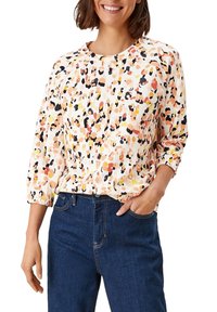 Bunt gemusterte Bluse in Orange, Gelb und Schwarz mit abstrakten Mustern, Rundhalsausschnitt und Dreiviertelärmel, kombiniert mit dunklen Jeans.