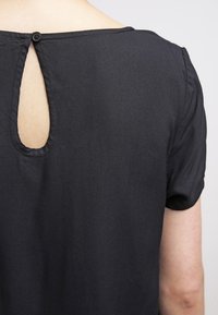 Vista posteriore di una persona che indossa una maglietta nera a maniche corte con una piccola apertura a forma di chiave fissata da un bottone nero vicino al collo.