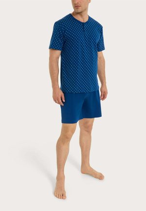 Uomo che indossa una camicia blu a maniche corte con motivo a puntini bianchi e pantaloncini blu uniti, in piedi a piedi nudi su uno sfondo chiaro.