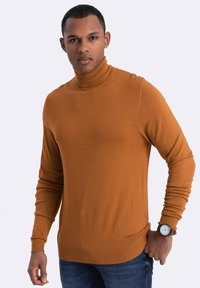 Turtleneck-Pullover in warmem Orange mit glatter Textur. Lange Ärmel und taillierte Passform. Getragen zu Denim-Jeans und einer Armbanduhr.