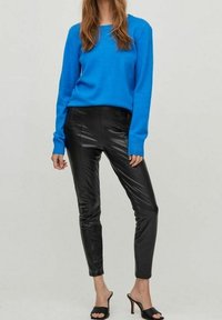 Mujer con un suéter de manga larga azul brillante, pantalones negros de cuero sintético y sandalias negras de tacón abierto, de pie contra un fondo claro.