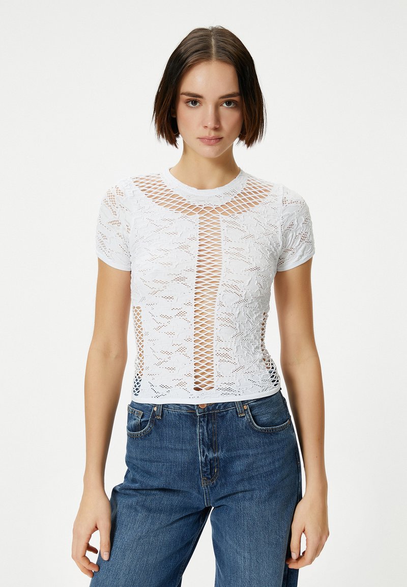 Witte kant t-shirt met een doorzichtige mesh ontwerp, ronde halslijn, korte mouwen en een centraal verticale patroon. Draagt over blauwe jeans.