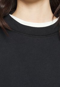 Sweat-shirt noir à texture côtelée et col rond, superposé à une chemise blanche. Présente un détail de couture au niveau du col.