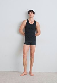 Schwarzes Tanktop und passende Shorts an einem männlichen Model, das mit den Händen hinter dem Rücken gegen eine graue Wand steht, barfuß auf einem hellen Teppich.