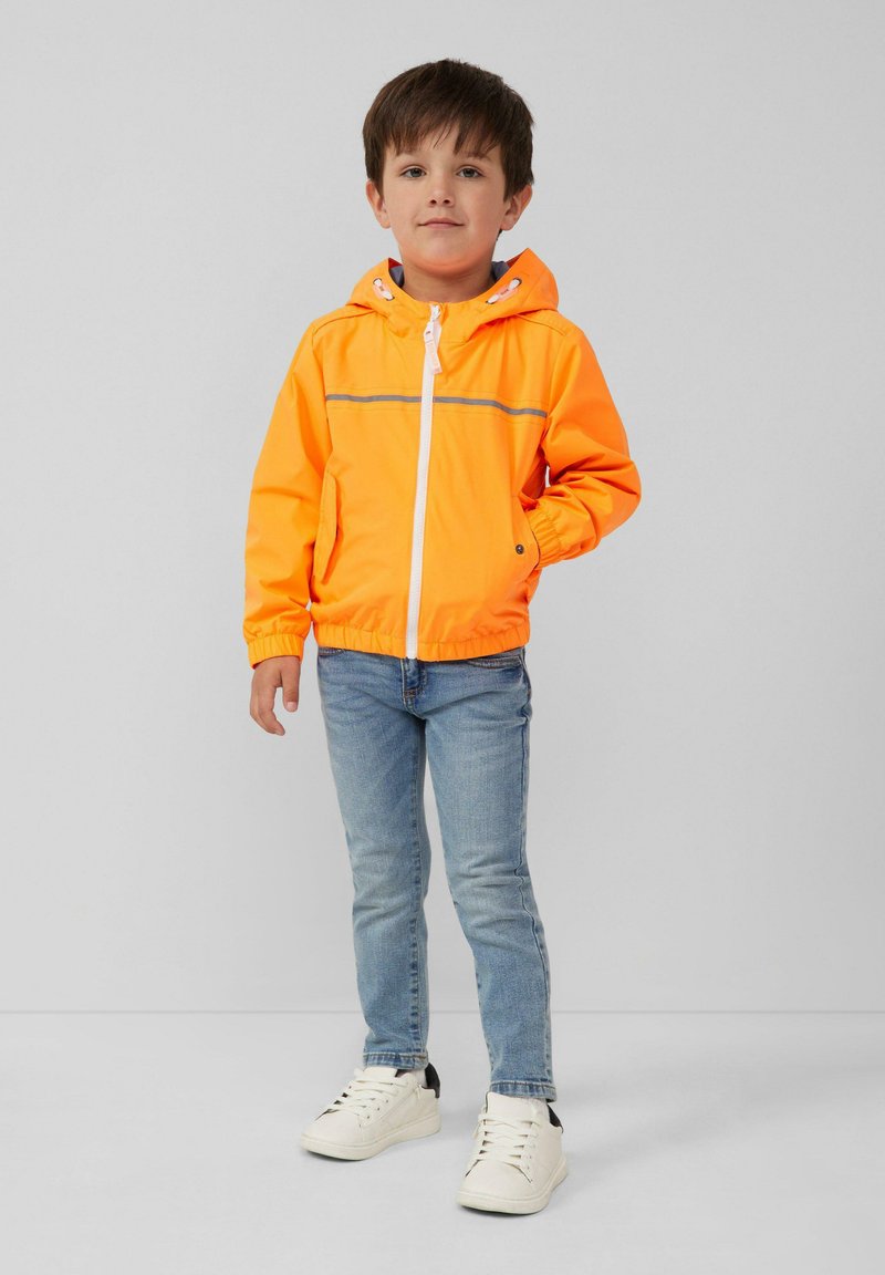 Heldere oranje lichtgewicht jas met capuchon, voorrits en twee zijzakken. Gepaard met blauwe jeans en witte sneakers.