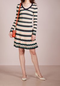 Klänning med lång ärm i en zickzackmönster i pastellrosa, grönt och svart. Har en rundad halsringning och är kombinerad med en röd axelväska.