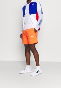 Chaqueta de entrenamiento Adidas en azul, rojo y blanco; pantalones cortos naranjas; y zapatillas blancas con rayas negras y acentos neón.