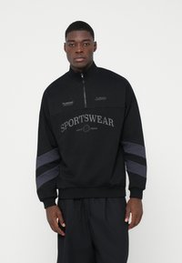 Giovane uomo in piedi che indossa una felpa sportiva nera con strisce a forma di chevron grigie sulle maniche e il testo "SPORTWEAR" sul petto.