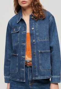 Chaqueta vaquera de azul profundo con cierre de botones en la parte delantera, dos bolsillos en el pecho y un corte relajado. Presenta un desgaste ligero y detalles en hardware plateado.