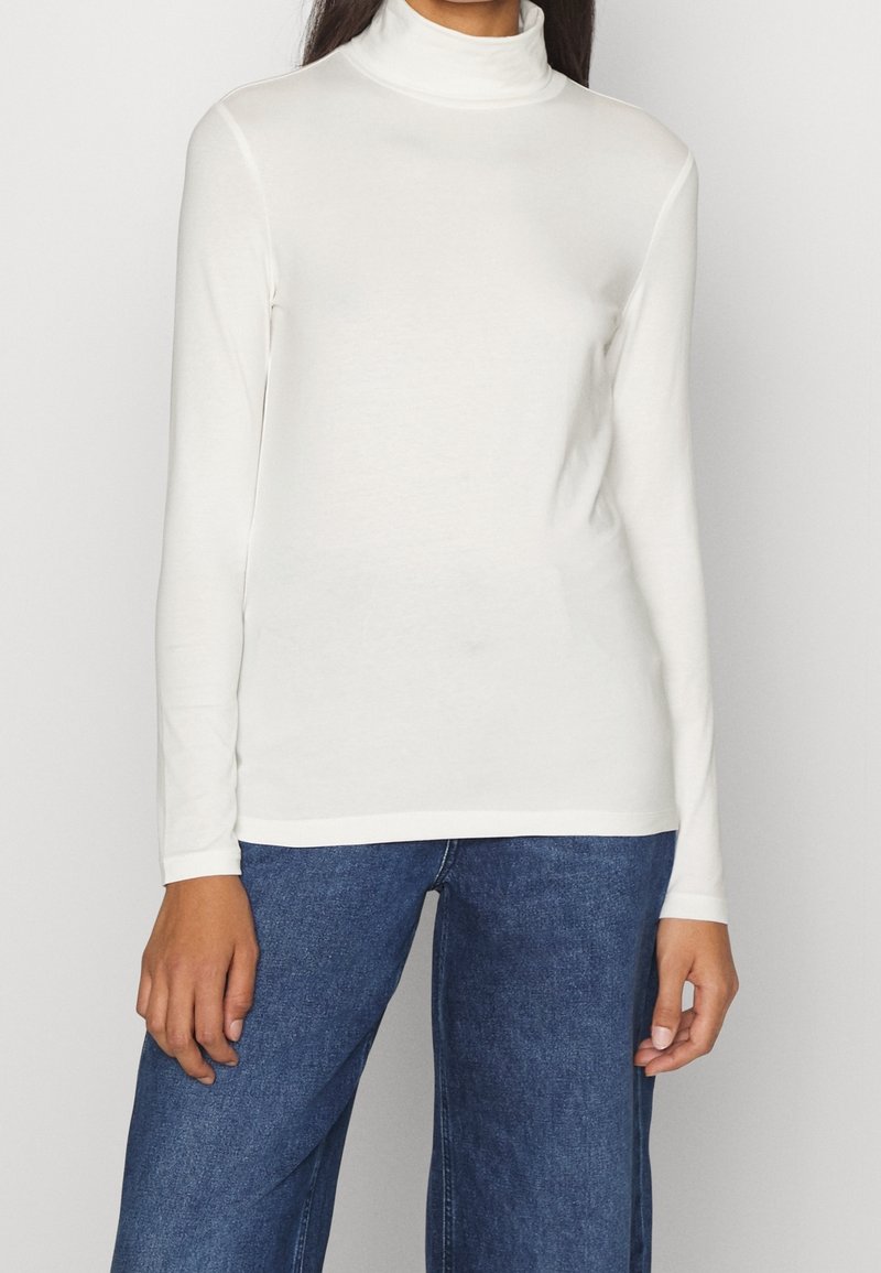 Weißes Langarm-Turtleneck-Oberteil mit glatter Textur, tailliertem Design und dezentem Elasthan-Anteil. Kombiniert mit dunkelblauen, weit geschnittenen Jeans.