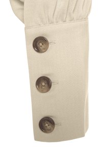 Beiger Stoffmanschette mit drei großen, dunkelbraunen Knöpfen. Die Manschette hat ein plissiertes Design und eine strukturierte Oberfläche.
